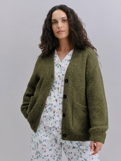 A woman wearing an olive green knit cardigan over a white pajama set with a colorful floral print, photographed against a solid light gray studio background.