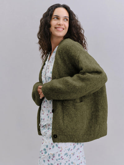 A woman wearing an olive green knit cardigan over a white floral pajama set, photographed against a solid light gray studio background.
