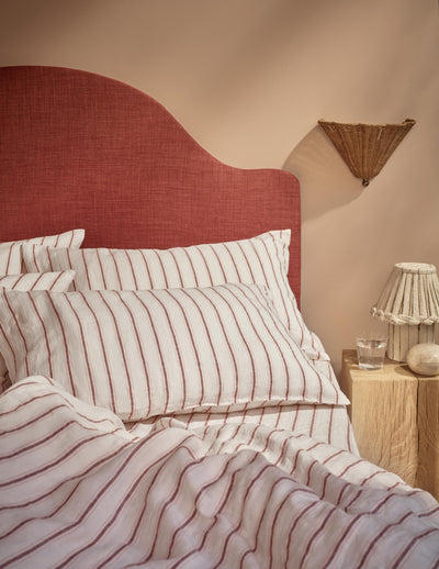 A styled bedroom scene showing a bed with white bedding featuring thin red vertical stripes, a curved red upholstered headboard, and a warm beige wall. A wooden bedside table with a woven lampshade lamp and a glass is visible on the right.