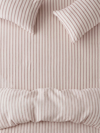 Close-up of a striped bedding set with a duvet cover and two pillows in a soft blush pink color with thin vertical dark red stripes.