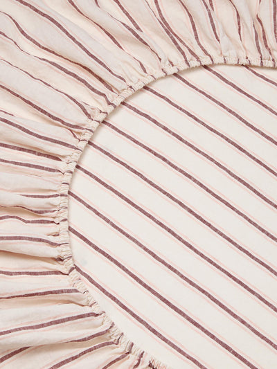 A close-up of a striped fitted sheet, featuring diagonal stripes in dusty rose and cream with a gathered ruffled edge.