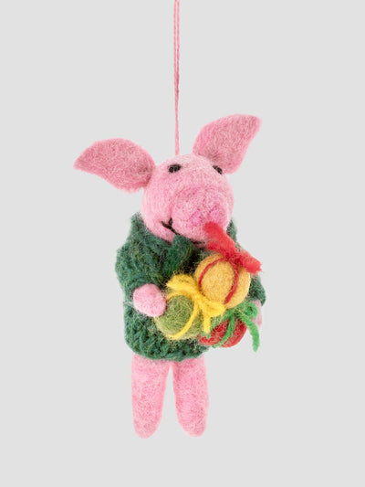 A felted wool Piglet Christmas ornament wearing a green sweater and holding a yellow and red flower, suspended by a thin pink string against a solid light gray background.
