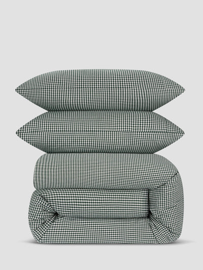 A stack of three bedding pieces with a small-scale green and white gingham check pattern, including two pillows and a folded comforter or duvet, set against a solid light gray background.