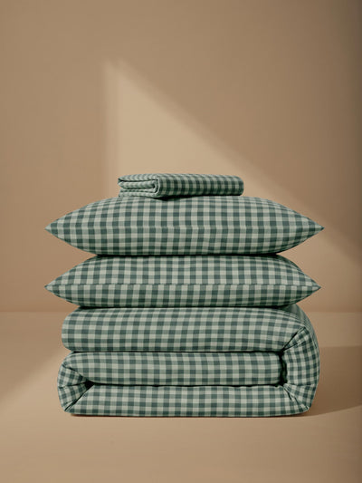A stack of four folded bedding items, including two pillows and two folded blankets, in a green and white gingham check pattern, set against a warm beige background with a soft shadow.