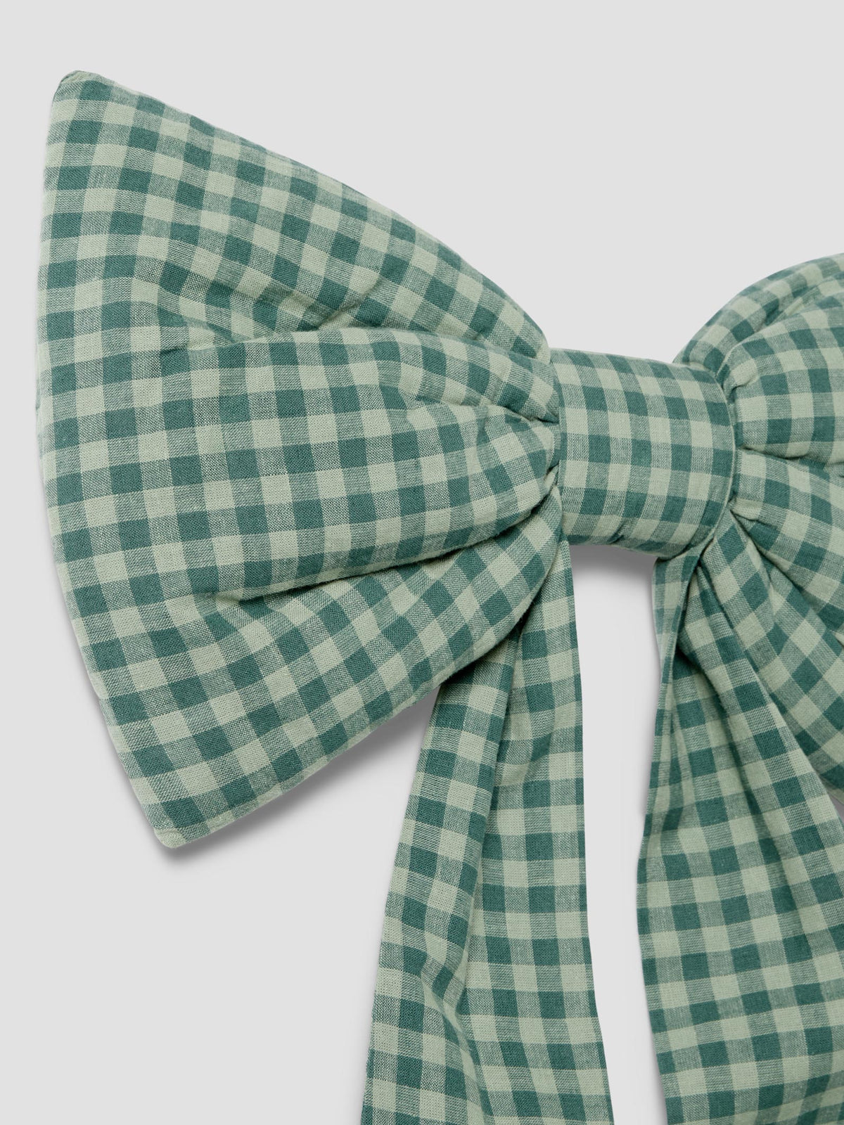 A close-up of a large bow tie made from green and off-white gingham fabric, set against a solid light gray background.