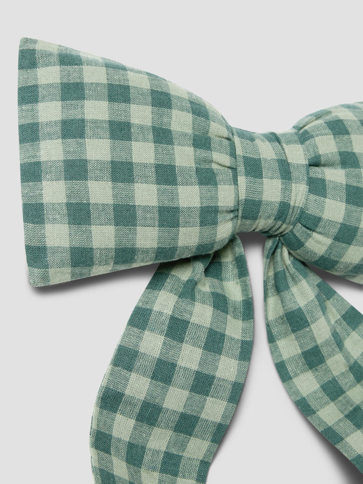 A close-up of a fabric bow in a green and off-white gingham check pattern, set against a solid light gray background.