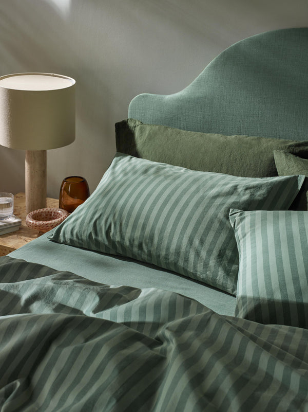 A styled bed with sage green and white striped bedding, including pillows and duvet, set against a muted green upholstered headboard in a softly lit bedroom. A bedside lamp and small amber glass bottle sit on the nightstand.