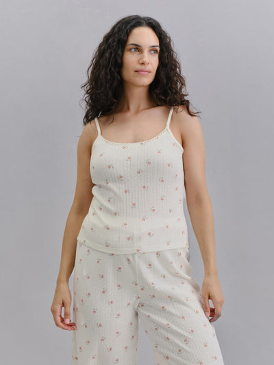 A woman modeling a two-piece ribbed pajama set in off-white with a small pink floral print, consisting of a camisole top and matching pants, against a solid gray background.