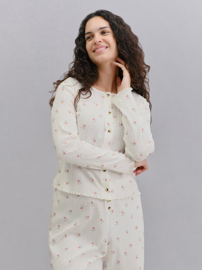A woman modeling a two-piece pajama set featuring a long-sleeve button-up top and matching pants in a cream-colored textured fabric with a small pink floral print, set against a solid gray background.