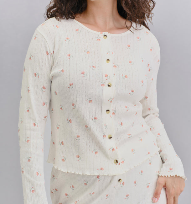 A woman wearing a cream-colored ribbed knit cardigan with a small pink floral print and gold buttons, paired with a matching skirt, against a solid gray background.
