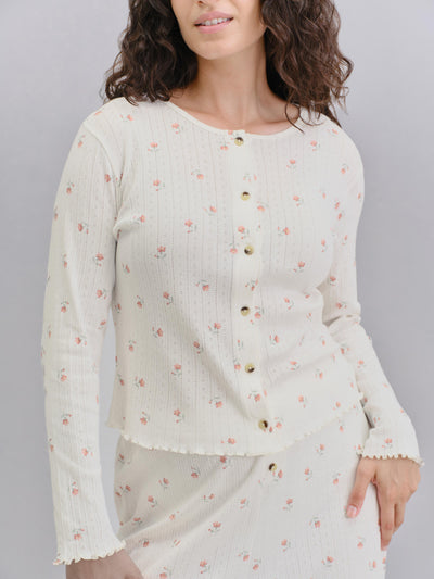 A woman wearing a cream-colored ribbed knit cardigan with a small pink floral print and gold buttons, paired with a matching skirt, against a solid gray background.