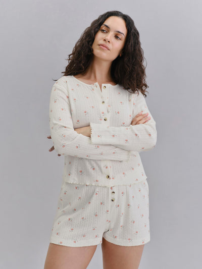 A woman modeling a matching two-piece ribbed knit loungewear set in off-white with a small pink floral print. The set includes a long-sleeve henley top and matching shorts, featuring gold-tone buttons.