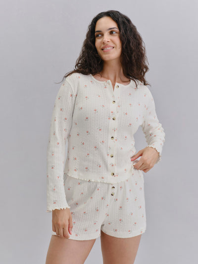 A woman modeling a two-piece ribbed knit loungewear set in off-white with a small pink floral print, featuring a long-sleeve button-front top and matching shorts, against a solid gray studio background.