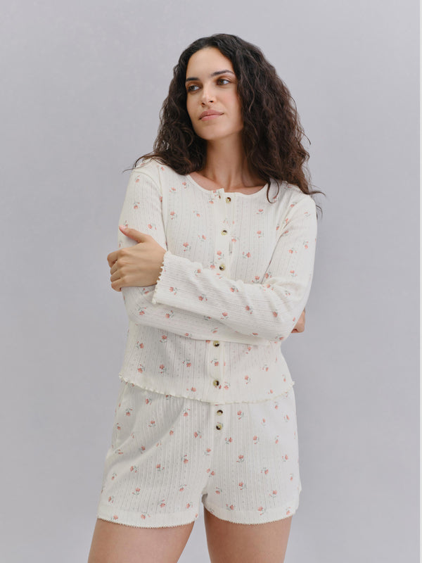 A woman modeling a two-piece pajama set consisting of a long-sleeve button-front top and matching shorts, both in an off-white/cream color with a small pink floral pattern, set against a plain light gray studio background.