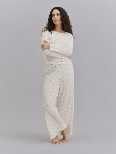 A woman modeling a two-piece long-sleeve pajama set in cream with a small pink floral pattern, standing against a solid gray studio background.