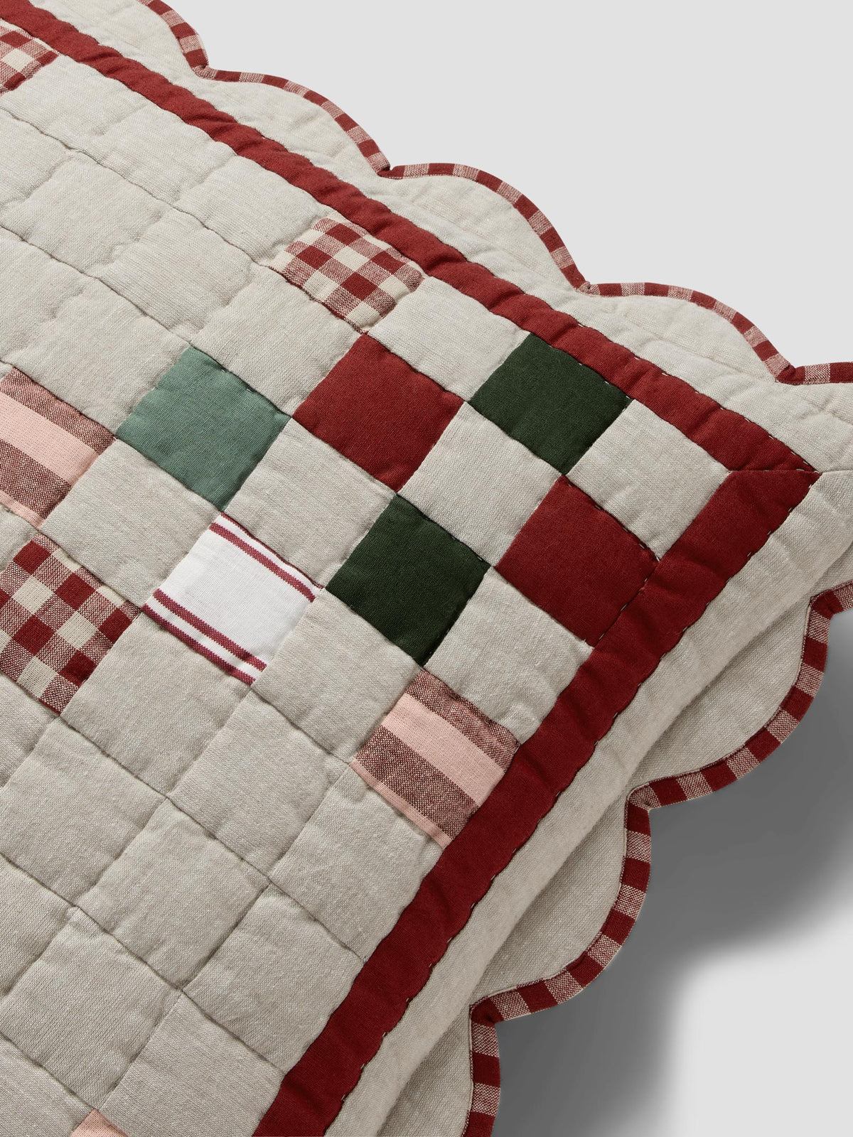 A close-up of a quilted patchwork pillow or cushion corner, featuring cream fabric, red gingham, red and green checkered squares, and a scalloped red border on a light gray background.
