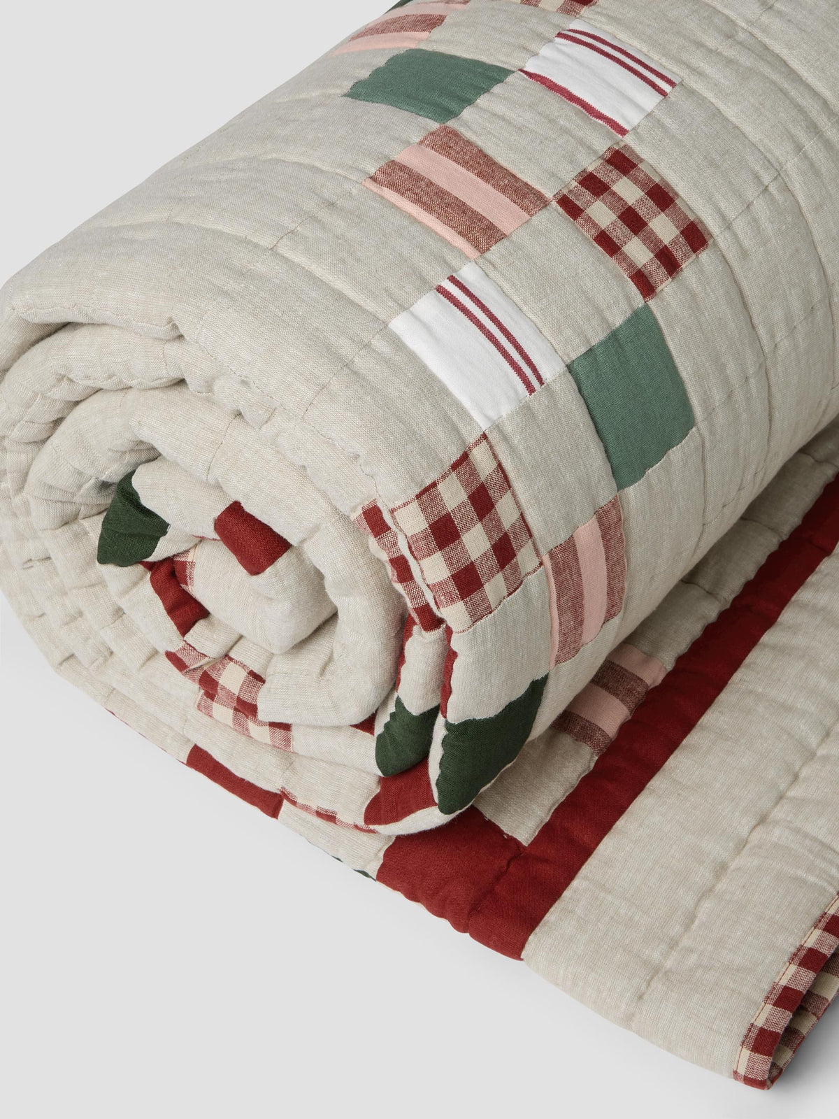 Rolled up beige patchwork blanket, folded on a light grey background. The blanket has a red piped edge, and green, red and navy blue patterned patch details. 