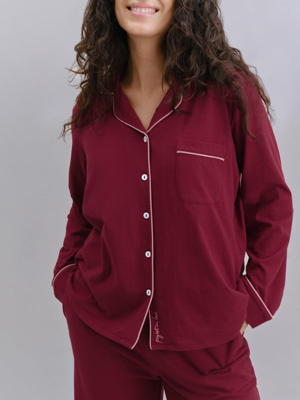 A woman wearing a burgundy long-sleeve pajama shirt with white piping and matching pajama pants, shown against a plain gray studio background.