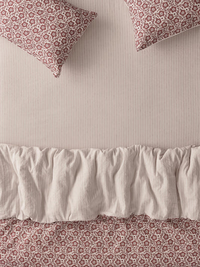 A close-up of bedding featuring a cream ruffled bed skirt and matching floral-patterned pillows and duvet in a muted dusty rose and cream print, set against a ribbed cream background.