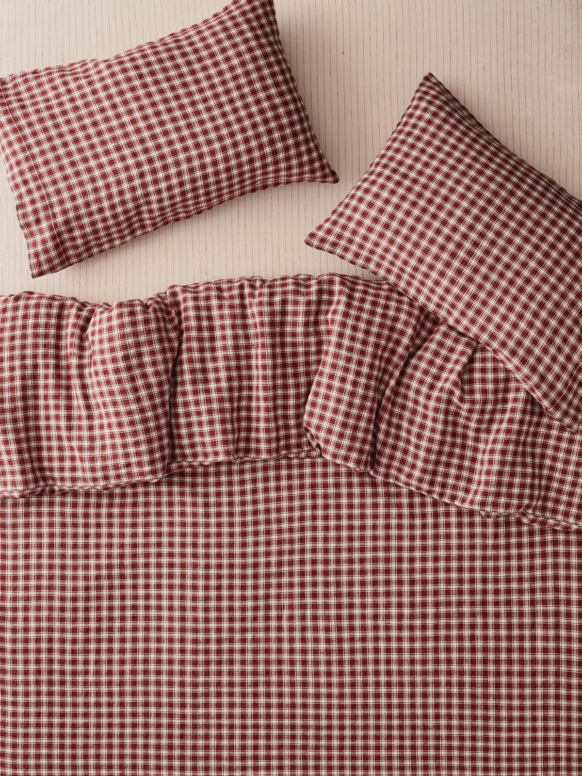A bedding set with a red and white gingham check pattern, including a duvet cover, a ruffled bedspread, and two matching pillowcases, arranged on a light beige textured background.