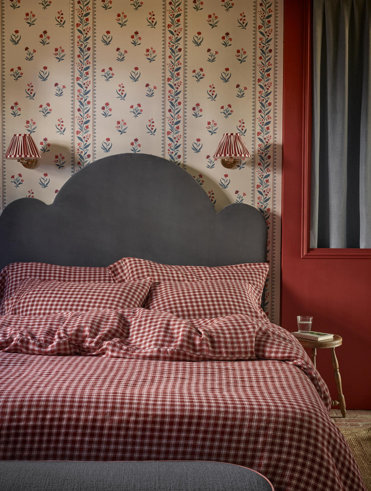 A styled bedroom scene featuring a bed dressed in red and white gingham check bedding, set against a cream floral wallpaper and a dark gray scalloped headboard. A dark red wall, wall sconces, and a small side table are visible.