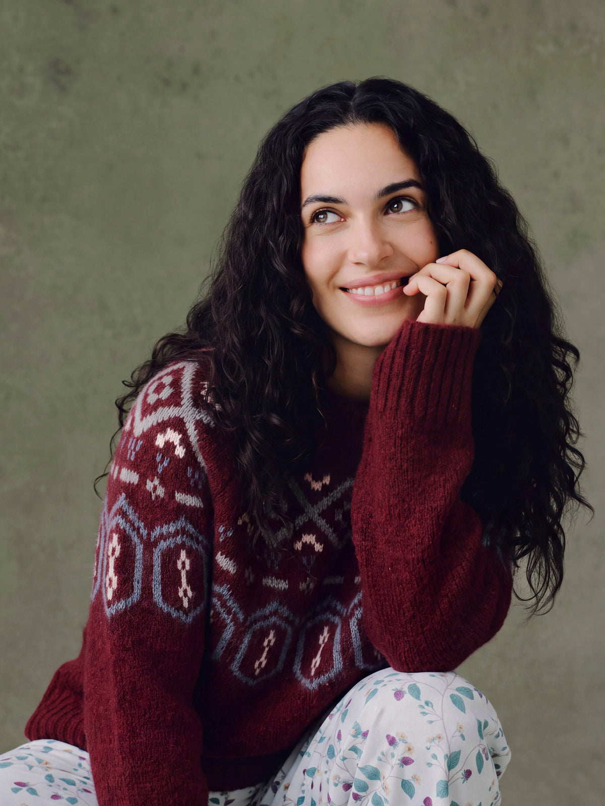 A woman with dark curly hair wearing a chunky knit burgundy sweater with a light blue and white geometric Fair Isle pattern across the chest and sleeves, posing against a textured sage green background.