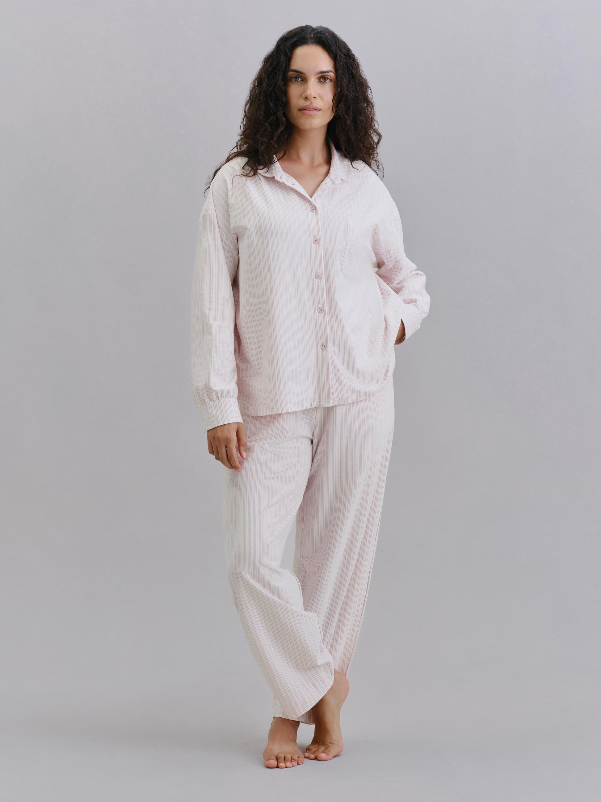 A woman modeling a light pink two-piece pajama set with subtle vertical stripes, consisting of a long-sleeve button-up shirt and matching pants, against a solid gray studio background.