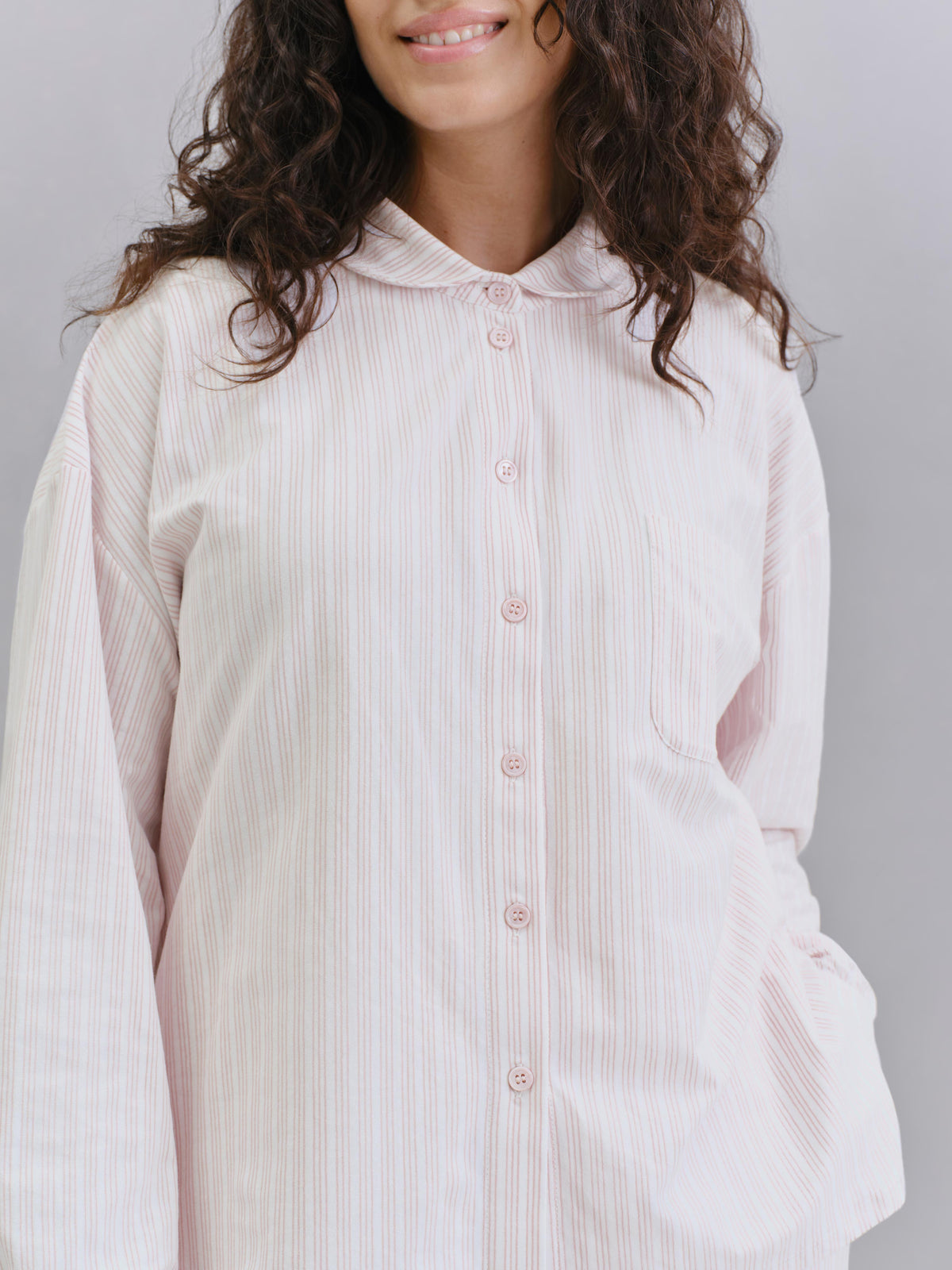 A woman wearing a pale pink and white vertically striped button-up shirt with a collar, shown from the chest up against a light gray studio background.