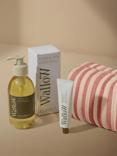 Wallow bath and body set featuring a pump bottle of Eternal Tea bath and body oil, a white box, a white tube, and a pink and white striped bath towel on a beige background.