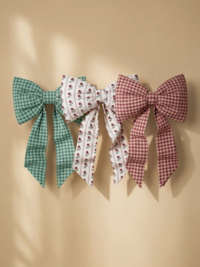 Three fabric hair bows in different patterns hang against a warm beige wall: a green and white gingham bow, a white bow with small red floral prints, and a dusty red and white gingham bow.