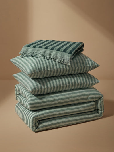 A stack of folded bedding pieces, including pillows and a folded blanket with fringe, in a muted sage green and white stripe pattern, set against a warm beige background.