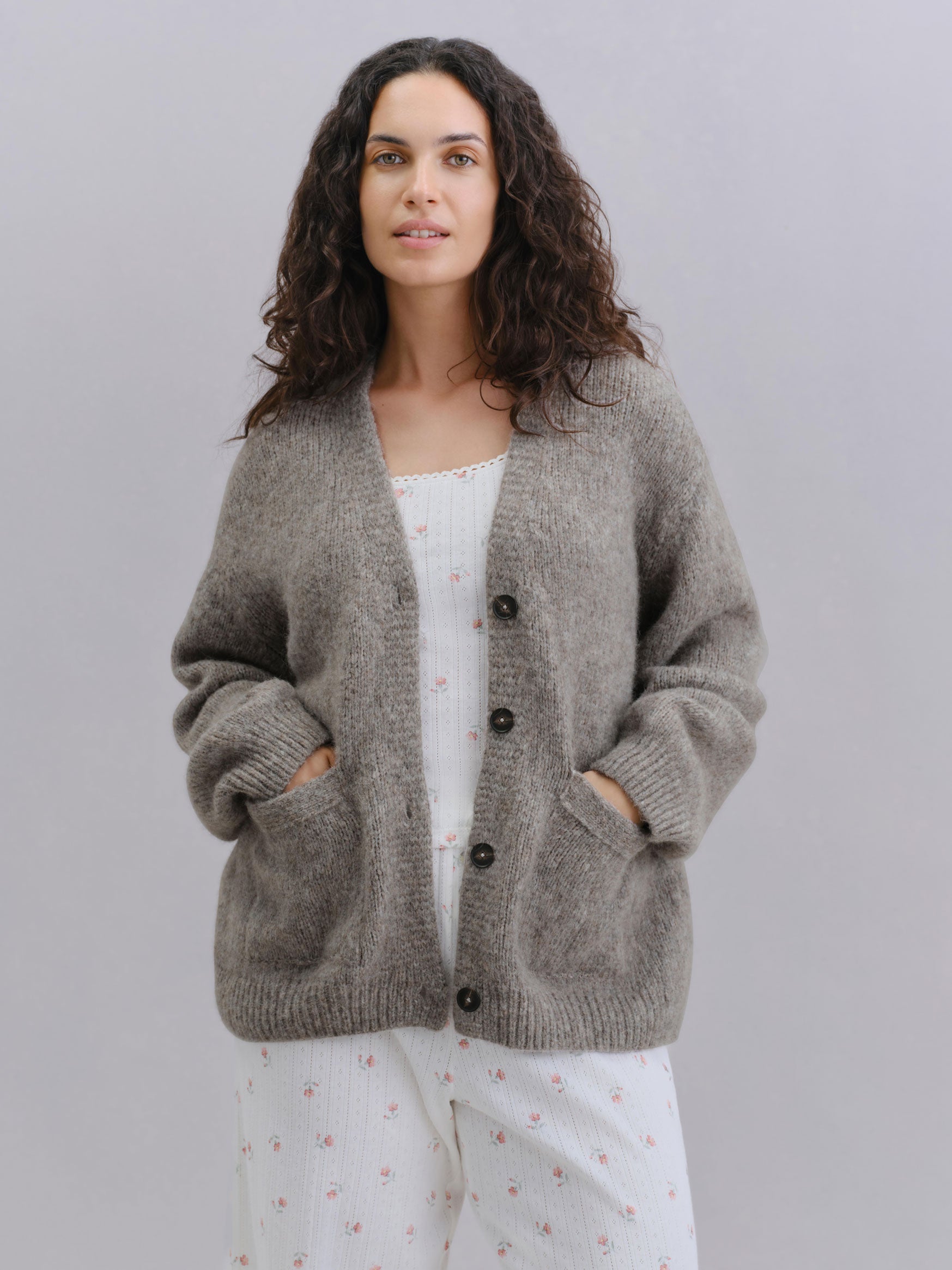 A woman with long, wavy dark brown hair is wearing a fuzzy, dark grey knit cardigan with front buttons and side pockets, layered over a white top and white pants with a small pink floral pattern. She is standing against a solid light gray studio background.