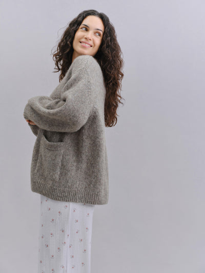A woman with long curly brown hair models an oversized, fuzzy taupe-gray knit sweater with a front patch pocket, paired with white patterned pants, against a solid light gray background.