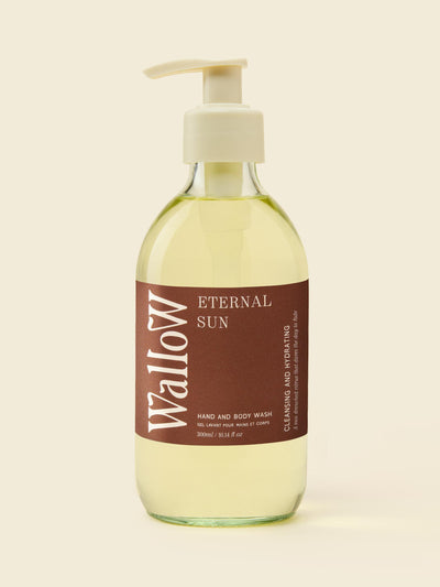 A bottle of Wallow Hand and Body Wash with a pump dispenser, filled with a pale yellow liquid, featuring a brown label with white text, set against a solid cream background.