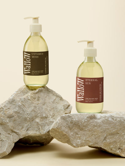 Two pump bottles of Wallow body wash, one labeled 'Untamed Moss' and the other 'Eternal Sun,' standing on large gray stones against a warm beige background.