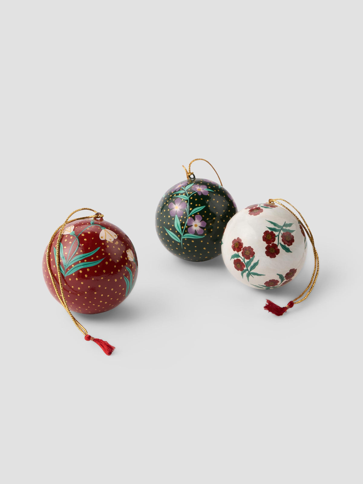 Three hand-painted papier mache Christmas ornaments with gold hanging loops, displayed on a light gray background. One ornament is dark red with gold dots and green leaves, one is black with gold dots and pink flowers, and one is white with red flowers and green leaves.