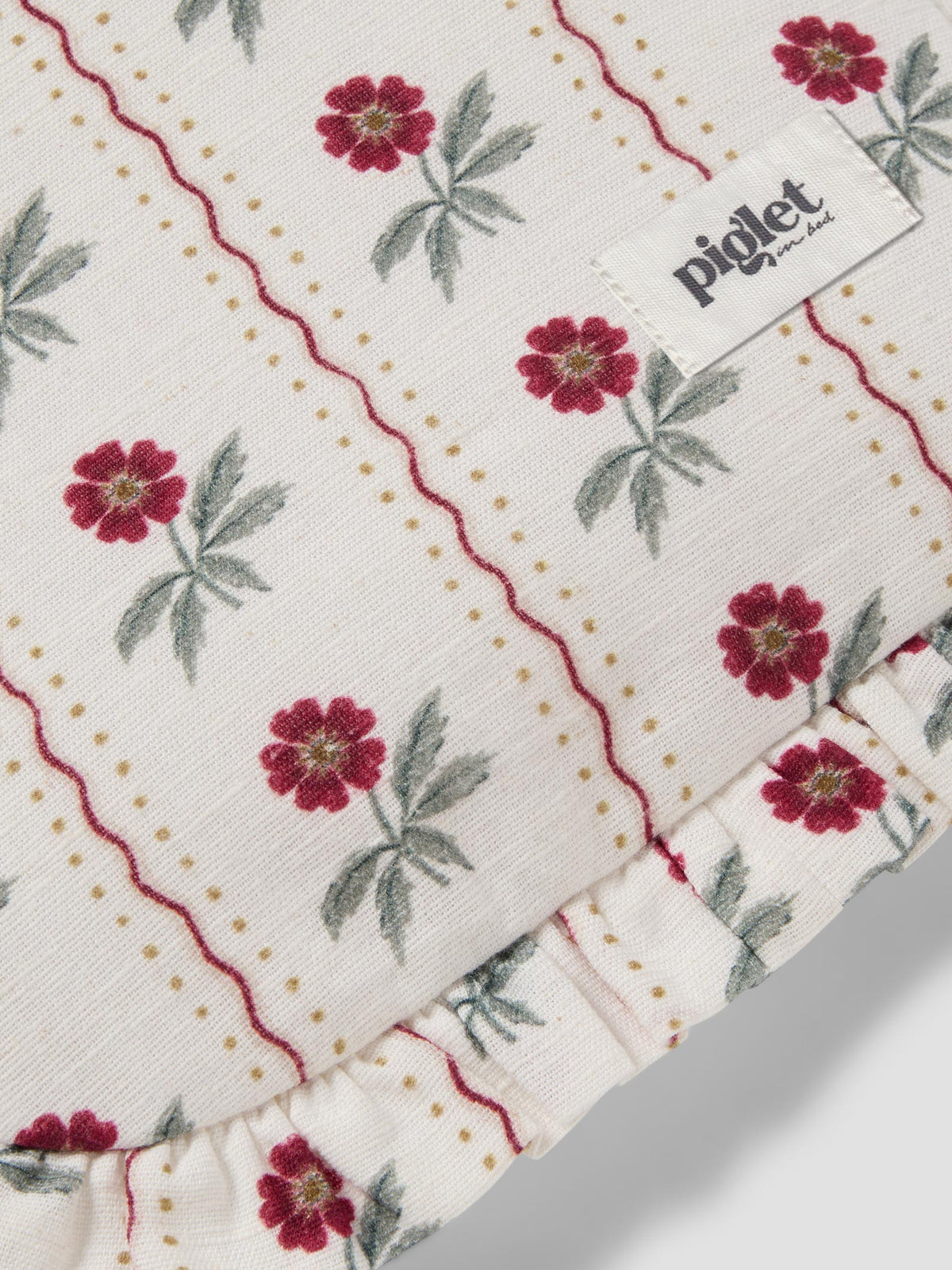Close-up of a cotton muslin textile with a cream background, featuring a red and green floral pattern, red wavy stripes, dotted lines, and a ruffled edge. A white brand label reading 'piglet' is visible.