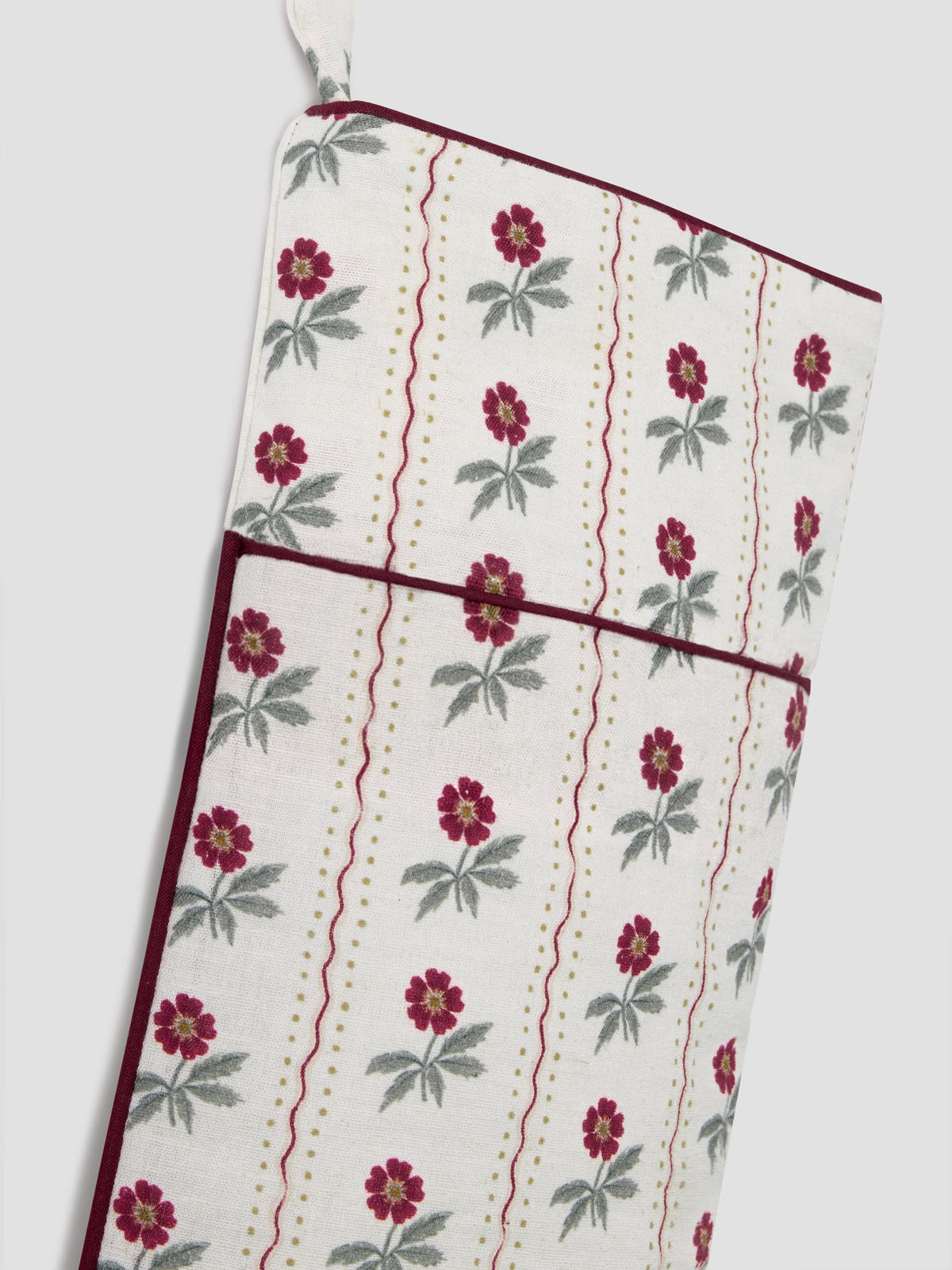 A close-up of a hanging stocking with a cream background, featuring a repeating pattern of red flowers with green leaves, vertical wavy red lines, and gold dotted stripes, finished with dark red piping.