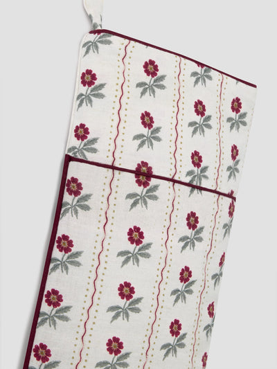 A close-up of a hanging stocking with a cream background, featuring a repeating pattern of red flowers with green leaves, vertical wavy red lines, and gold dotted stripes, finished with dark red piping.