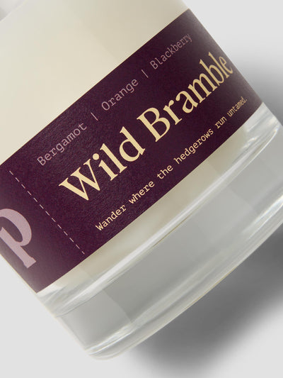 Close-up of a candle in a clear glass jar with a dark plum label reading 'Wild Bramble' and smaller scent text including Bergamot, Orange, and Blackberry.