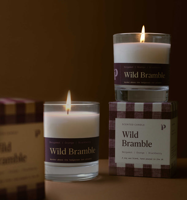 Three Wild Bramble scented candles in clear glass jars with white wax and dark purple labels, arranged on a warm brown surface with their branded boxes. One candle is lit and placed in front, another is lit and stacked on top of a box, and a third box is partially visible on the left.