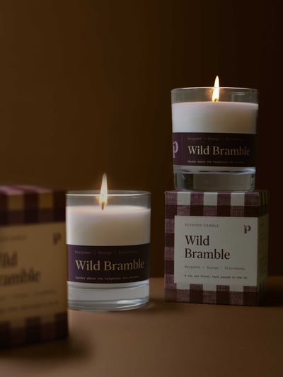 Three Wild Bramble scented candles in clear glass jars with white wax and dark purple labels, arranged on a warm brown surface with their branded boxes. One candle is lit and placed in front, another is lit and stacked on top of a box, and a third box is partially visible on the left.