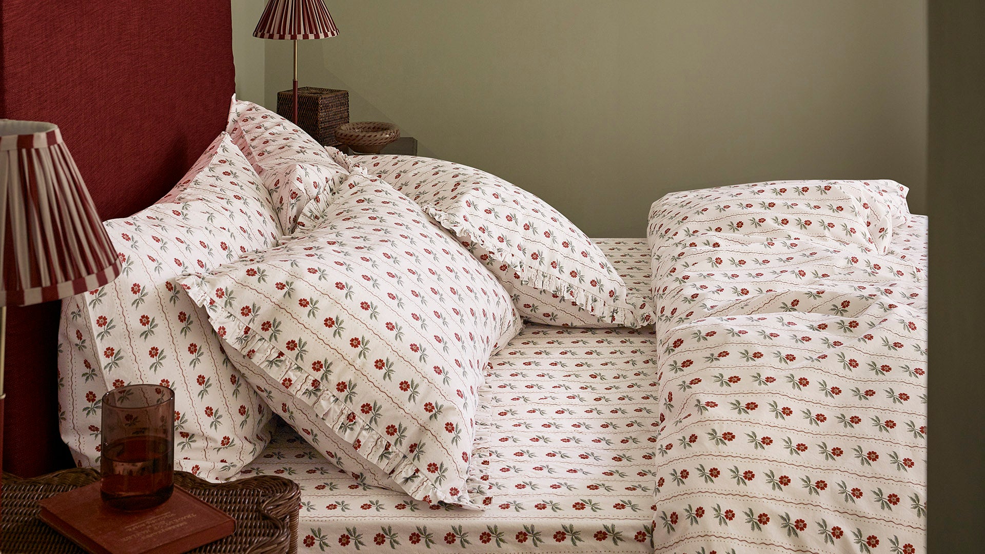 A made bed with white bedding printed all over with small red and green floral motifs, styled in a bedroom with matching pillows, a red headboard, and bedside lamps.